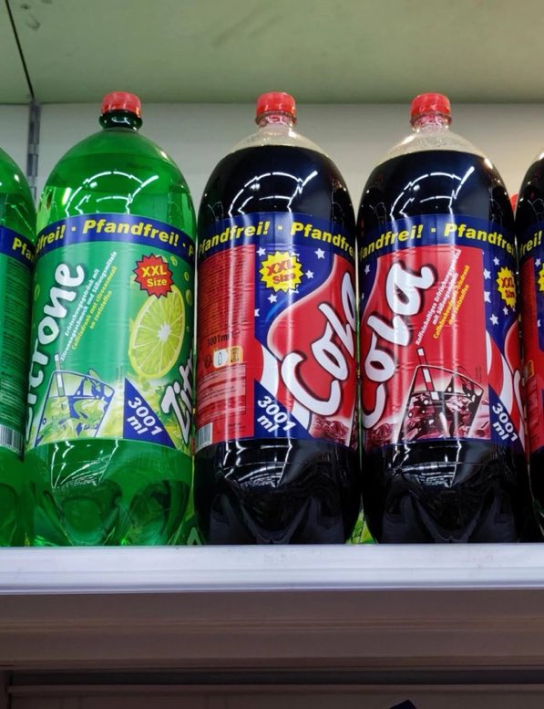Three large plastic bottles on a store shelf. The bottles are labeled Pfandfreii and contain 3,001 liters each, with flavors including lemon-lime and cola. The labels feature red, green, and blue colors with star designs.