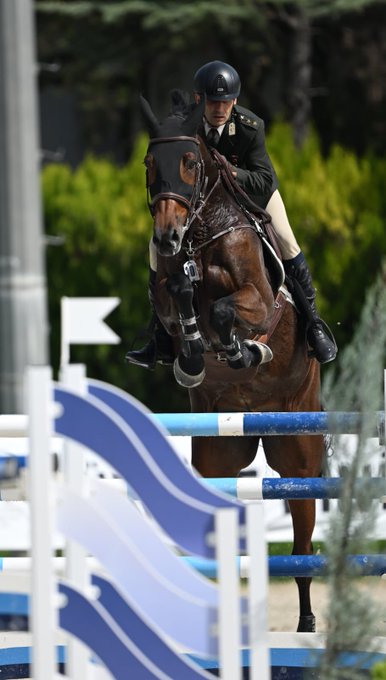 Burak Uluçay riding a horse named Kutlu, mid-jump over a blue and white obstacle in an equestrian event. He wears a helmet, dark jacket, and riding boots. A second image shows Burak Uluçay holding a trophy, surrounded by people in suits and uniforms, clapping. Banners display "Cumhurbaşkanlığı Kupası" and "Türkiye Büyük Millet Meclisi."