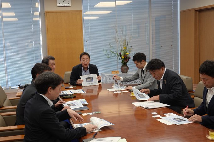 Yamazoe Taku, Renbi Sohei, and Otaira Yoshinobu sit at a wooden table in a meeting room with Fukuda Yoshihiko. They hold documents and discuss, with a floral arrangement and lace tablecloth visible. Blinds cover the windows, and cups are on the table. A nameplate reads "Fukuda."