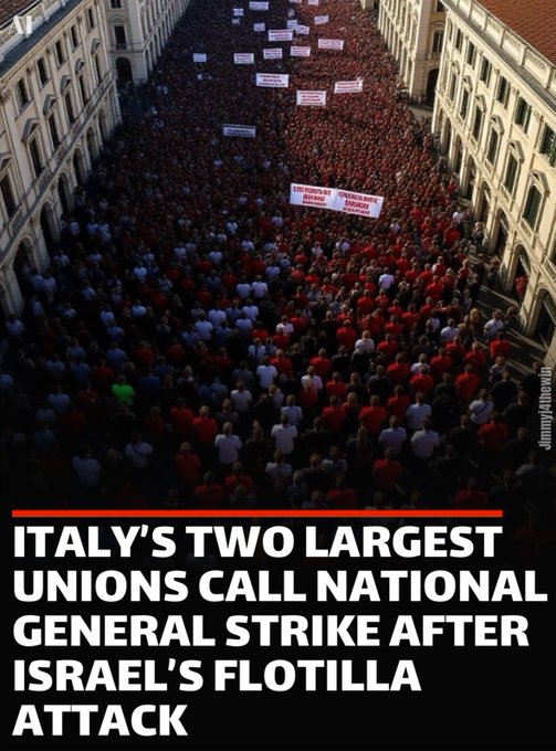 A large crowd of people gathered in a narrow urban street between tall buildings, holding red and white signs and banners. Some banners display text in Italian.