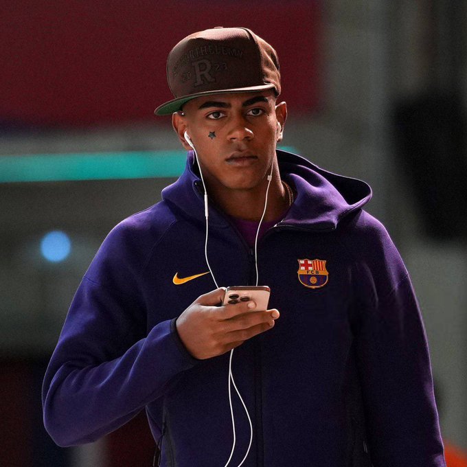Lamine Yamal wearing a purple FC Barcelona hoodie with the club logo, holding a smartphone with earphones plugged in, and wearing a brown cap with a visible "R" on it. A star is drawn on his face.