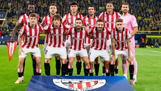 A group of soccer players wearing red and white striped jerseys, white shorts, and black socks, standing on a grassy field. They are positioned in two rows, with some kneeling and others standing, in front of a blue platform displaying the Athletic Club Bilbao logo. One player on the right wears a pink jersey.