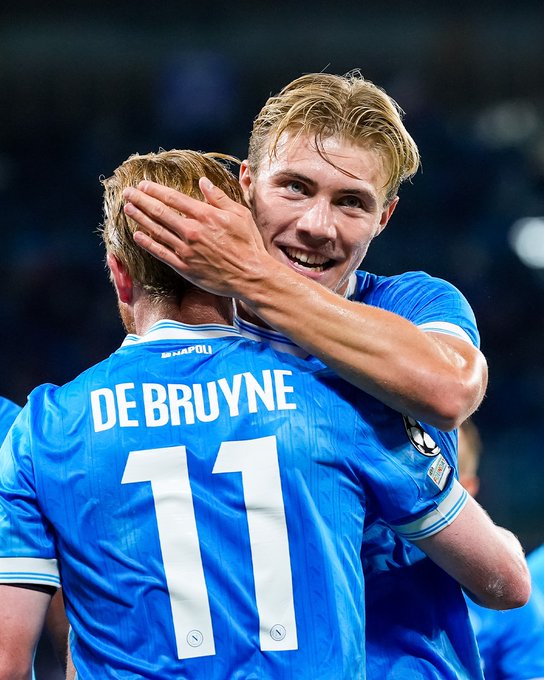 Rasmus Højlund and Kevin De Bruyne in blue soccer jerseys, embracing on a field. The jersey on the player in front has the name "DE BRUYNE" and the number 11. The background shows a stadium with blurred spectators.