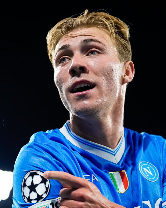Rasmus Højlund wearing a blue soccer jersey with the UEFA Champions League patch and the Napoli logo. He is pointing at the patch on his sleeve. The jersey features an Italian flag design.