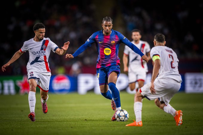 Alejandro Balde in a blue and red FC Barcelona jersey dribbling a soccer ball on a grassy field. He is surrounded by opposing players in white and red jerseys. The scene takes place during a match, with a stadium crowd visible in the background.