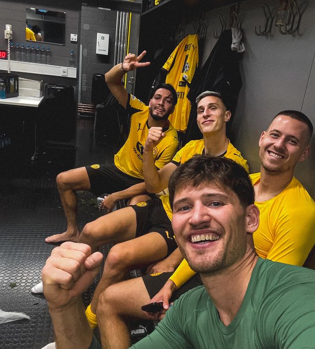 Four men in Borussia Dortmund yellow jerseys and black shorts, sitting and posing together in a locker room. One man in a green shirt sits in front, smiling and holding a phone for a selfie. The locker room has gray walls, black flooring, and hanging jerseys on hooks.