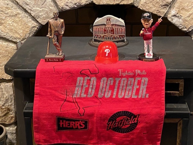 bloggerhythms's tweet image. My wood stove is ready for Saturday night. This is a rally towel from the game that Rhys Hoskins hit his famous bat-spike HR with a replica of the Harry Kalas statue. A Charlie Manuel bobblehead is on the right. In the background is a carboard cutout of Connie Mack stadium.