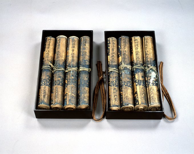 Two black rectangular boxes containing rolled scrolls tied with string. The scrolls have visible text and illustrations, including ink writing on the covers reading 