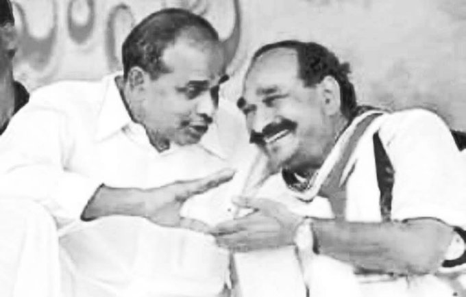 A black-and-white photo showing two men in conversation. Both are wearing white shirts, with one gesturing with his hand. The background includes a wall with circular patterns.