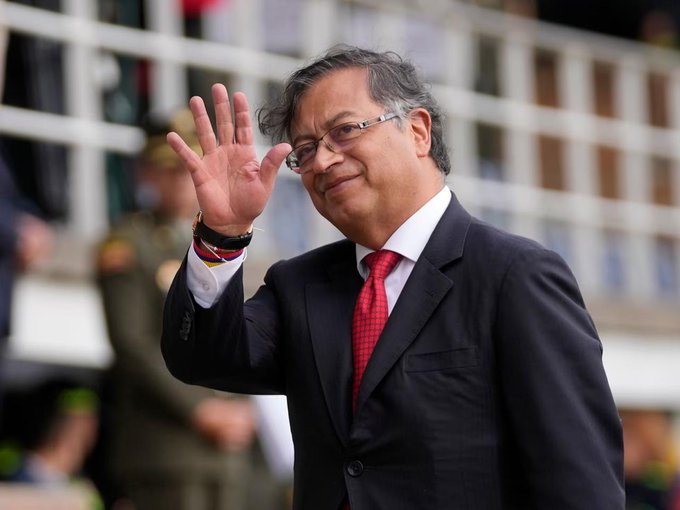 Gustavo Petro waving with his right hand raised, wearing a dark suit, white shirt, red tie, and a wristband with yellow, blue, and red colors. He stands outdoors near a building with white railings and other people in the background.