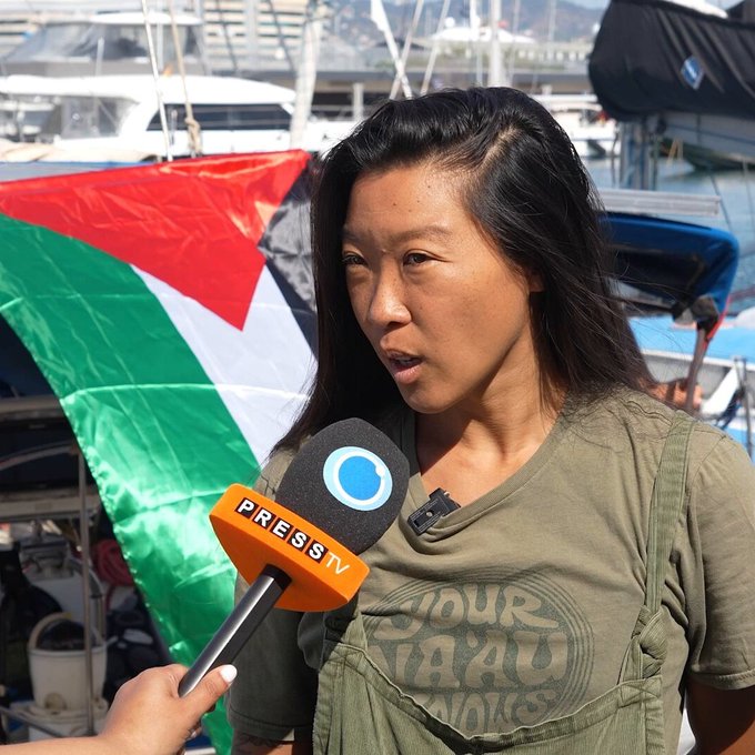 Jasmine Ikeda, a woman with long dark hair, wearing a green t-shirt and overalls, speaking into a microphone with a "PRESS" label. A Palestinian flag is visible in the background, along with boats and a marina setting.