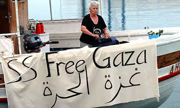 A boat docked at a Gaza port with people on board and on the dock. The boat displays Palestinian flags and a banner reading "SS Free Gaza" in English and Arabic. Another boat with Palestinian flags and people on deck is visible in the water.