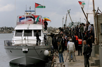 A boat docked at a Gaza port with people on board and on the dock. The boat displays Palestinian flags and a banner reading "SS Free Gaza" in English and Arabic. Another boat with Palestinian flags and people on deck is visible in the water.