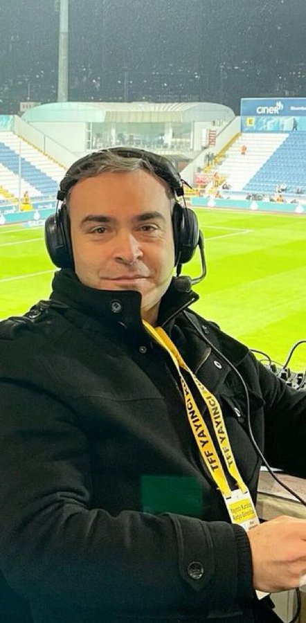 Onur Şahin wearing headphones and a black jacket, sitting in a stadium with a green field and stands visible in the background. A yellow lanyard with text hangs around his neck.