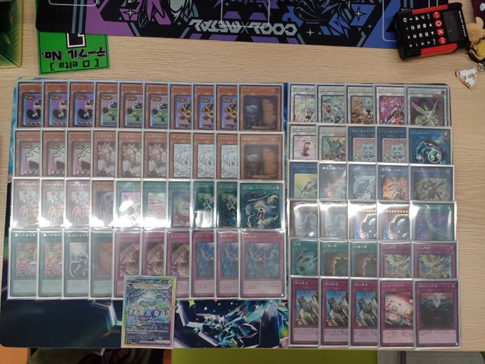 Multiple Yu-Gi-Oh trading card decks organized in binders and loose stacks on a desk. The cards feature colorful anime-style artwork of monsters and characters. A calculator, a playmat with abstract designs, and a small figurine are also visible on the desk.