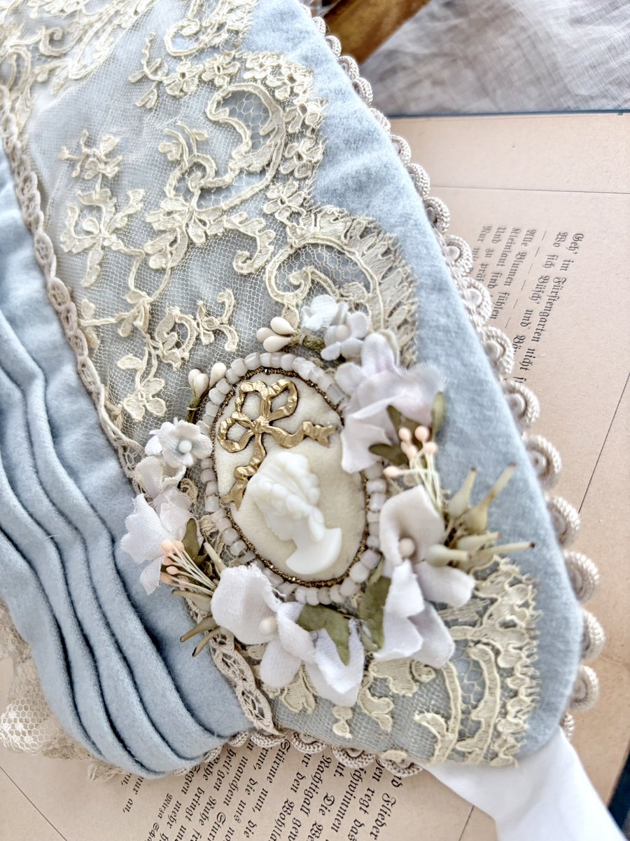 少しくすんだ、落ち着いた色味の水色のウールでハーフボンネットを制作しました。

ウール生地の上から繊細で美しいアンティークレースを被せています。
両サイドには陶器でできたカメオとメタルのリボン、それらを囲むようにビロードのすみれや勿忘草などのお花を縫い付けています。