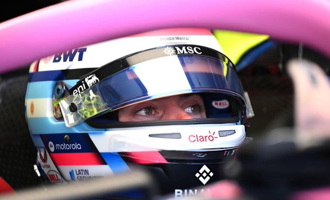 A close-up of a Formula 1 driver wearing a helmet with visible sponsors including BWT, Motorola, Claro, and Eni. The helmet features a white base with blue, red, and pink accents. The driver is seated in a race car, gripping the steering wheel, with part of the pink car visible.