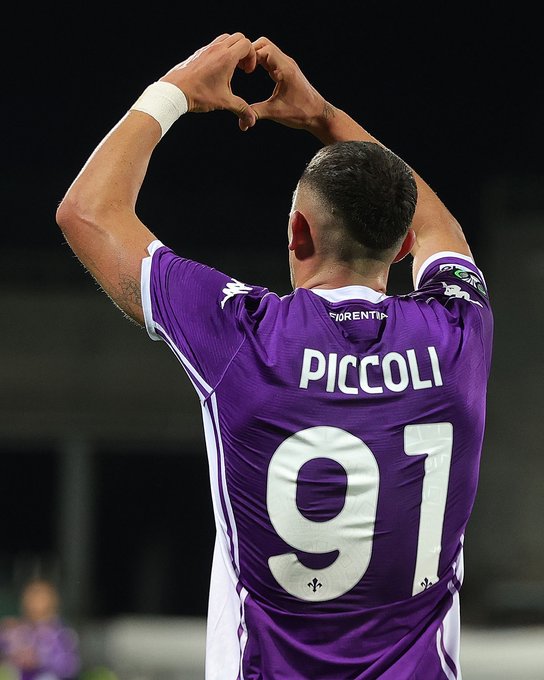 Lorenzo Piccoli wearing a purple soccer jersey with the name "PICCOLI" and number 91 on the back. He is making a heart shape with his hands above his head. The jersey has white accents and the word "FIORENTINA" visible on the upper back.