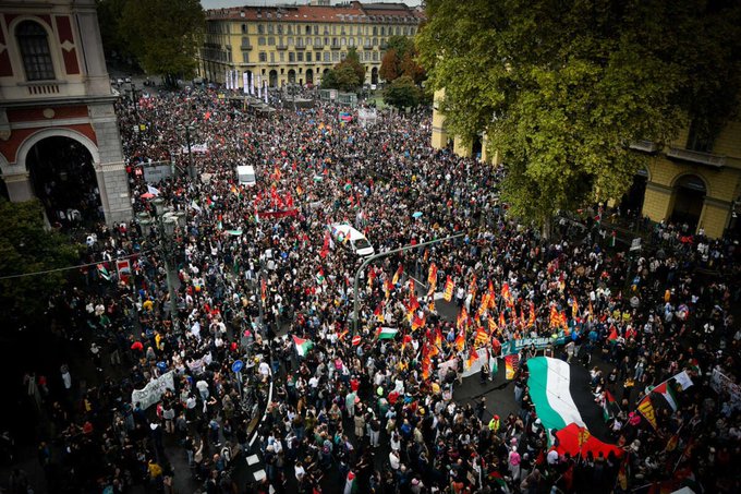A large crowd of people gathered in an urban street, holding signs and banners. Many carry Palestinian flags, visible among the sea of participants. The scene includes buildings and trees lining the street, with some vehicles visible in the distance.