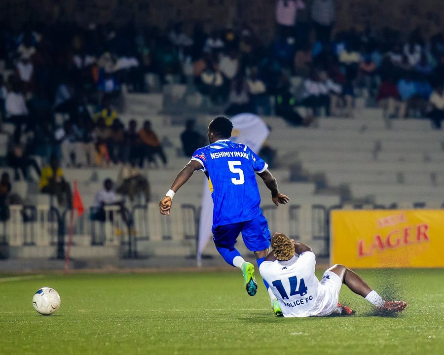 A soccer player in a blue jersey with "KIHWANGA" and the number 5 on the back, running on a grassy field. Another player in a white jersey with "MUJAWAMARIYA" and the number 14 on the back is on the ground. A crowd is visible in the stands, and a yellow advertisement banner with "LAGER" is on the sideline.