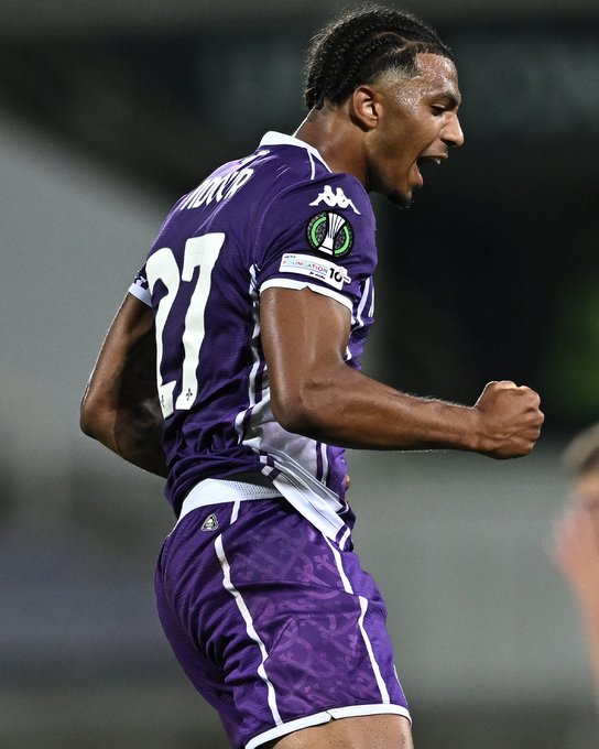 Cher Ndour wearing a purple soccer jersey with the number 27 and the name "Ndour" on the back. He is celebrating with a clenched fist and an open mouth, appearing to shout. The jersey features a logo on the sleeve and a badge on the chest.