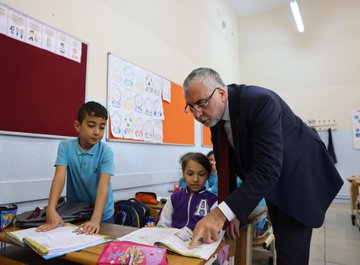 A group of students in blue uniforms holding colorful balls stand on steps in front of a pink building with the text "VALİ KAZI PAŞA İLK OKULU" above the entrance. Inside a classroom, students in blue shirts sit at desks with open books, and a man in a suit interacts with them, pointing at a book. Bulletin boards with drawings and educational materials are visible on the walls.