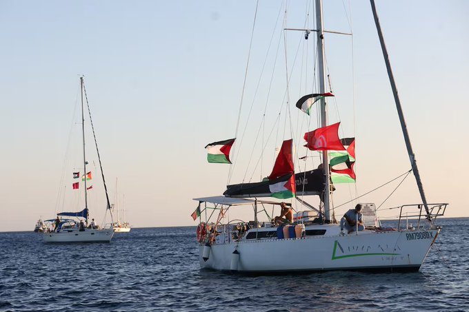 Two sailboats on the ocean. The closer boat, Marinette, is white with multiple Palestinian flags and red flags visible on its mast and deck. Several people are on board. The second boat in the background has various flags, including a Palestinian flag. The name "Marinette" is visible on the hull of the closer boat.