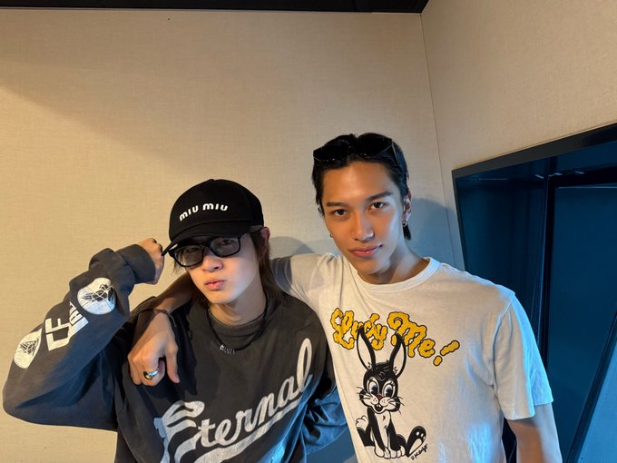 Rito Ikezaki wearing a black cap with "miu miu" text and a gray sweatshirt with "Eternal" text, standing next to Yudai Sano in a white T-shirt featuring a cartoon bunny and "Lucky Me!" text. Both are indoors, with Rito Ikezaki posing with an arm raised and Yudai Sano standing close, both smiling.