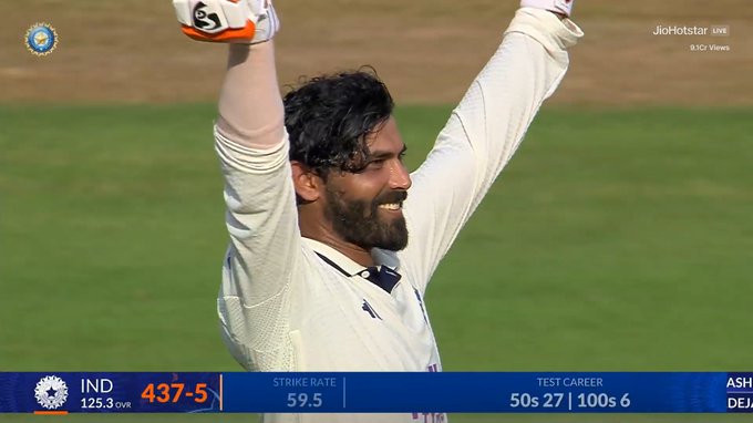 Ravindra Jadeja on a cricket field, wearing a white uniform with long sleeves and gloves, raising both arms in celebration. A scoreboard shows 