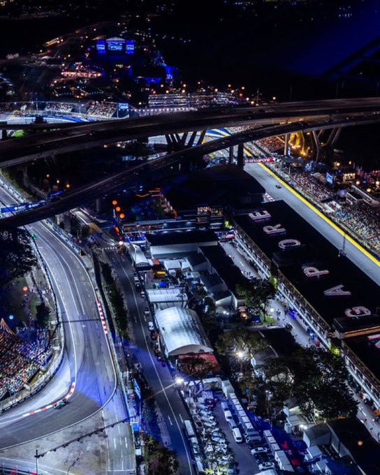 An aerial view of a brightly lit race track at night, featuring multiple lanes and overpasses. Cars are visible on the track, illuminated by lights. The track includes a section with the word "SINGAPORE" visible on a building alongside the circuit.