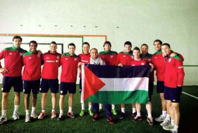 A group of men wearing red and green AthleticClub uniforms standing on a grassy field indoors, holding a Palestinian flag. They are positioned closely together, some with arms around each other, wearing matching shorts and socks.