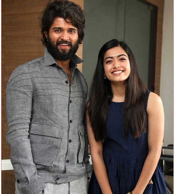 Vijay Deverakonda wearing a grey buttoned shirt and light trousers, standing next to Rashmika Mandanna in a sleeveless navy blue dress. Both are smiling, with Vijay Deverakonda having a beard and long hair, and Rashmika Mandanna with long straight hair.