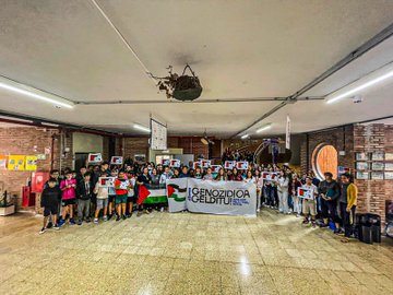 A group of people standing in a line outdoors in a town square, holding a large banner reading "IKASLEOK GENOZIDIA GELDITU PALESTINAREKIN" with Palestinian flags. Some individuals hold smaller signs with text and flags. Nearby, another group poses in a parking lot holding a similar banner and flags, with buildings and trees visible. A third group stands indoors on a tiled floor, holding banners and flags, with brick walls and posters in the background.