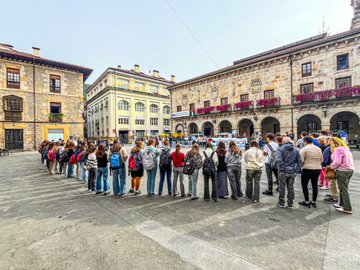 A group of people standing in a line outdoors in a town square, holding a large banner reading "IKASLEOK GENOZIDIA GELDITU PALESTINAREKIN" with Palestinian flags. Some individuals hold smaller signs with text and flags. Nearby, another group poses in a parking lot holding a similar banner and flags, with buildings and trees visible. A third group stands indoors on a tiled floor, holding banners and flags, with brick walls and posters in the background.