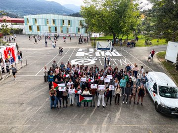 A group of people standing in a line outdoors in a town square, holding a large banner reading "IKASLEOK GENOZIDIA GELDITU PALESTINAREKIN" with Palestinian flags. Some individuals hold smaller signs with text and flags. Nearby, another group poses in a parking lot holding a similar banner and flags, with buildings and trees visible. A third group stands indoors on a tiled floor, holding banners and flags, with brick walls and posters in the background.