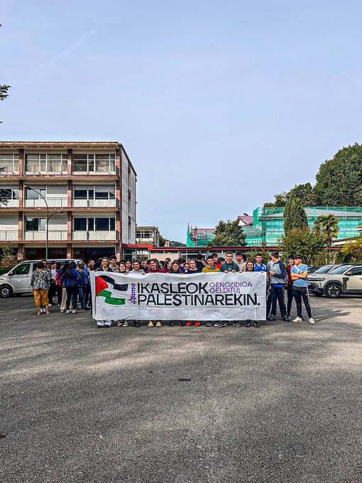 A group of people standing in a line outdoors in a town square, holding a large banner reading "IKASLEOK GENOZIDIA GELDITU PALESTINAREKIN" with Palestinian flags. Some individuals hold smaller signs with text and flags. Nearby, another group poses in a parking lot holding a similar banner and flags, with buildings and trees visible. A third group stands indoors on a tiled floor, holding banners and flags, with brick walls and posters in the background.