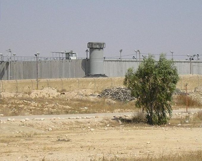 A concrete wall with a guard tower and barbed wire, surrounded by a barren desert landscape. A small tree and scattered rocks are visible in the foreground. The wall and tower are part of Ktzi\'ot prison in the Néguev Desert.