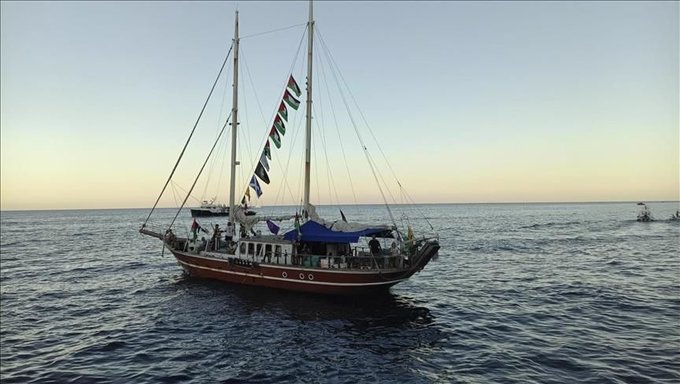 A large sailboat with two masts on the open sea, decorated with colorful flags along the rigging. The vessel has a blue canopy on deck and appears to carry people. Another smaller boat is visible in the distance on the water.