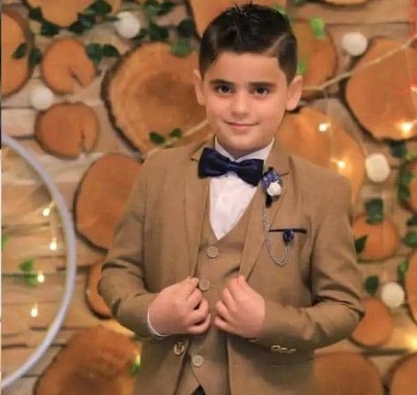 A young boy with dark hair, wearing a tan suit, white shirt, and blue bow tie, standing with hands clasped. A blue pocket square and a flower boutonniere are visible on his suit. The background features wooden slices, green leaves, and white pom-poms with string lights.