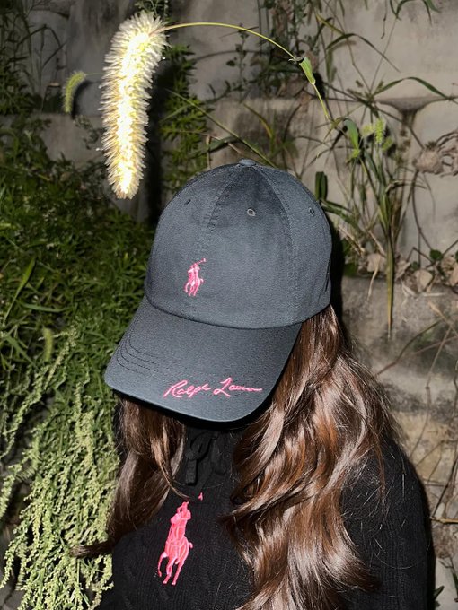 Krystal Jung wearing a black hoodie with a pink Polo Ralph Lauren logo on the chest, paired with black pants and a black handbag. Long brown hair flows over her shoulders. She stands in front of a green leafy plant and a white textured wall in one image, and in front of a similar wall with a potted tree in another. A third image shows her wearing a black Polo Ralph Lauren cap with a pink logo, standing in front of greenery.