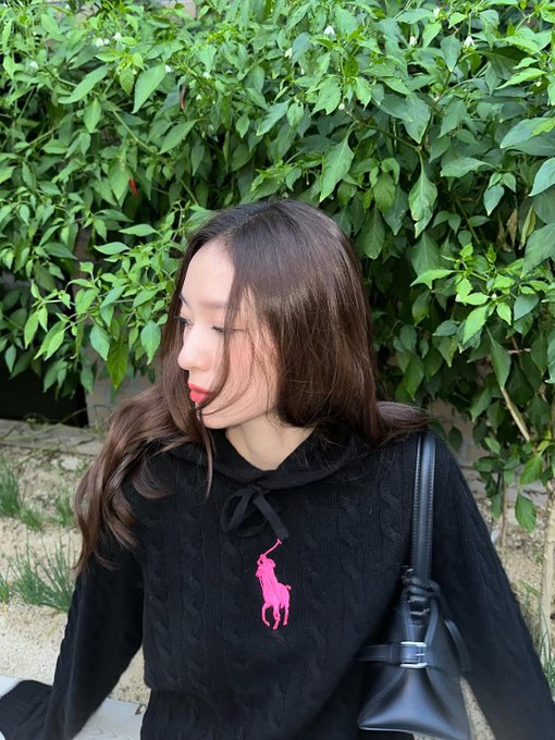 Krystal Jung wearing a black hoodie with a pink Polo Ralph Lauren logo on the chest, paired with black pants and a black handbag. Long brown hair flows over her shoulders. She stands in front of a green leafy plant and a white textured wall in one image, and in front of a similar wall with a potted tree in another. A third image shows her wearing a black Polo Ralph Lauren cap with a pink logo, standing in front of greenery.