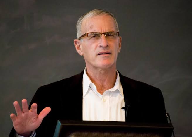 Norman Finkelstein standing at a podium, wearing glasses, a white shirt, and a dark blazer, gesturing with his right hand. A flag of Israel with a red prohibition symbol over it, featuring a diagonal line crossing out the Star of David.