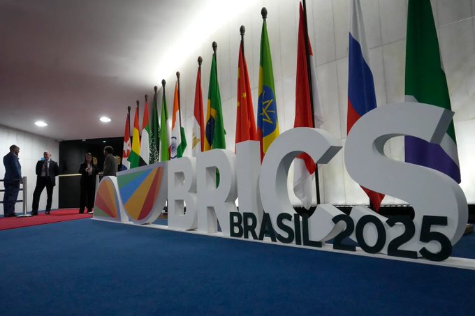 Vladimir Putin sitting at a table, wearing a dark suit and red tie, with hands clasped. Behind him are multiple Russian flags with blue, white, and red stripes. A separate image shows a display with the word "BRICS" in large letters, "BRASIL 2025" below, and flags of various countries including Russia, Brazil, and others.
