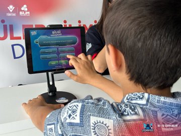First image displays several children at a table in a science festival setting, engaged in assembling a cardboard model resembling a vehicle or device, surrounded by paper sheets, glass slides, and molecular structure diagrams on a banner with Turkish text for TUBITAK and science education, event date October 10-12 2025. Second image shows a boy in a blue shirt touching a touchscreen quiz game interface displaying questions in Turkish about knowledge, with a banner in the background promoting science and technology education. Third image captures adults and a child at a booth holding and displaying white t-shirts printed with red science-themed designs, seated around tables with laptops and chairs under a tent-like structure at the outdoor festival. Fourth image features two young boys, one wearing a VR headset holding a purple device and the other grasping a paper airplane, both in casual clothing with one in a blue and claret soccer jersey, standing near a table in the event area with blurred background elements.