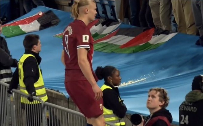 Erling Haaland in red soccer jersey stands on blue stadium tarp holding green white black red Palestinian flag with white triangle several security guards in yellow vests and black clothing around him diverse group including women and men some wearing casual clothes others in uniforms blue tarp covers seating area with scattered flags and banners in background.
