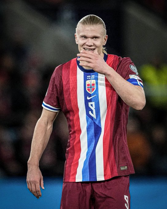 Erling Haaland stands on a soccer field wearing a red white and blue Norwegian national team jersey with the number 9 on his back and a sponsor patch on the sleeve his blonde hair in a ponytail he smiles widely covering his mouth with his right hand raised in celebration the background shows stadium seating with blurred spectators and a blue sky.