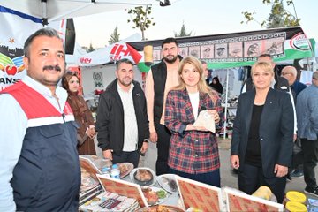 First image shows two women, one in a black headscarf and long dress standing next to a younger woman in a plaid skirt and white blouse, with large posters of a man with white hair and beard in the background under a tent with Turkish and Palestinian flags, fabrics hanging, and chairs around. Second image depicts a group of people including men in vests and women in headscarves and blouses standing near a tent with Palestinian flags and banners, tables displaying food like plates of meat and drinks, books, and promotional items. Third image features several women, some wearing headscarves in various colors, conversing near a green tent with hanging clothes, bags, toys, and fabrics on display tables. Fourth image shows people including a woman in plaid blouse and another in headscarf browsing stalls under a large green banner reading Gazze için Genel Kıyafet Yardımı with images of children, tents with clothes and aid items displayed.