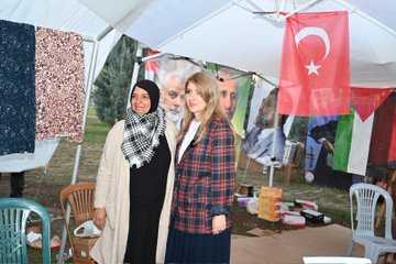 First image shows two women, one in a black headscarf and long dress standing next to a younger woman in a plaid skirt and white blouse, with large posters of a man with white hair and beard in the background under a tent with Turkish and Palestinian flags, fabrics hanging, and chairs around. Second image depicts a group of people including men in vests and women in headscarves and blouses standing near a tent with Palestinian flags and banners, tables displaying food like plates of meat and drinks, books, and promotional items. Third image features several women, some wearing headscarves in various colors, conversing near a green tent with hanging clothes, bags, toys, and fabrics on display tables. Fourth image shows people including a woman in plaid blouse and another in headscarf browsing stalls under a large green banner reading Gazze için Genel Kıyafet Yardımı with images of children, tents with clothes and aid items displayed.
