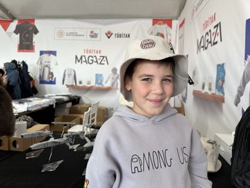 First image shows a young boy in a gray Among Us hoodie smiling at a TÜBİTAK Mağaza booth with shelves of books, t-shirts, and boxes under a tent with TÜBİTAK logos and banners. Second image depicts a woman in a red headscarf handing a white mug to a boy while a man takes a photo, surrounded by books, more mugs, and people at the booth. Third image features two girls examining white mugs with red splash designs and Turkish text on a table with books. Fourth image shows a man holding up a white t-shirt with red splash print and Turkish text, next to a woman and child, with stacks of clothing and products on display.