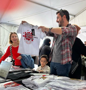 First image shows a young boy in a gray Among Us hoodie smiling at a TÜBİTAK Mağaza booth with shelves of books, t-shirts, and boxes under a tent with TÜBİTAK logos and banners. Second image depicts a woman in a red headscarf handing a white mug to a boy while a man takes a photo, surrounded by books, more mugs, and people at the booth. Third image features two girls examining white mugs with red splash designs and Turkish text on a table with books. Fourth image shows a man holding up a white t-shirt with red splash print and Turkish text, next to a woman and child, with stacks of clothing and products on display.